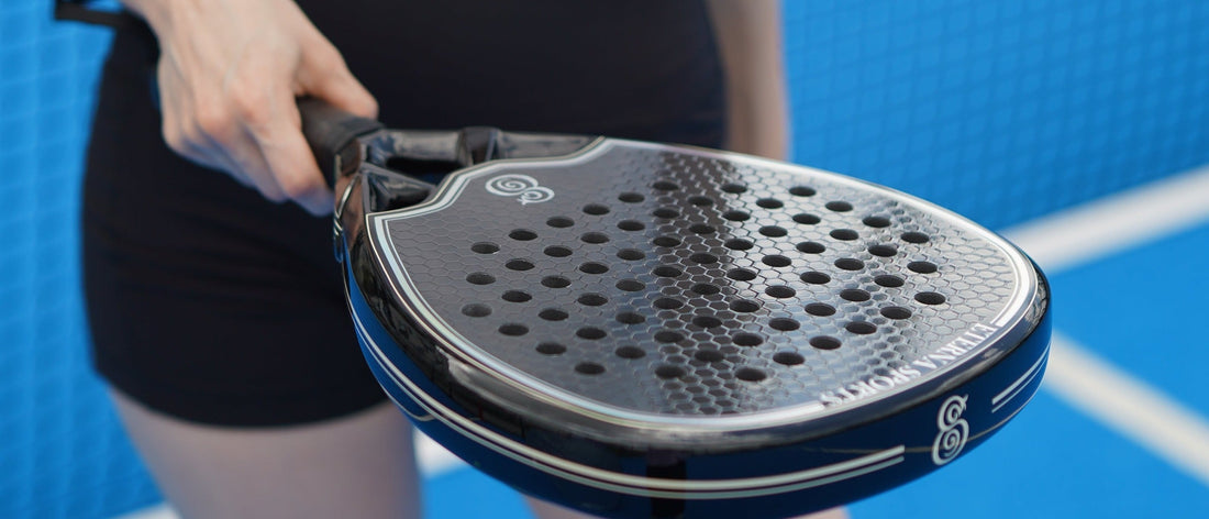 A hand on the grip of an Eterna padel racket, held level on a blue court