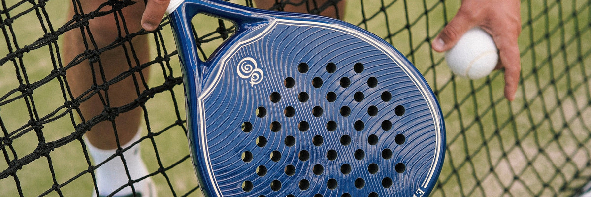 The face of an Eterna Soho Blue Glossy padel racket held over the net on a green court, a ball in the other hand